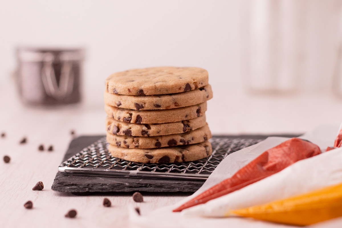 stack of 6 chocolate chip cookie cutouts on a cooling rack with 3 bags of royal icing next to it.