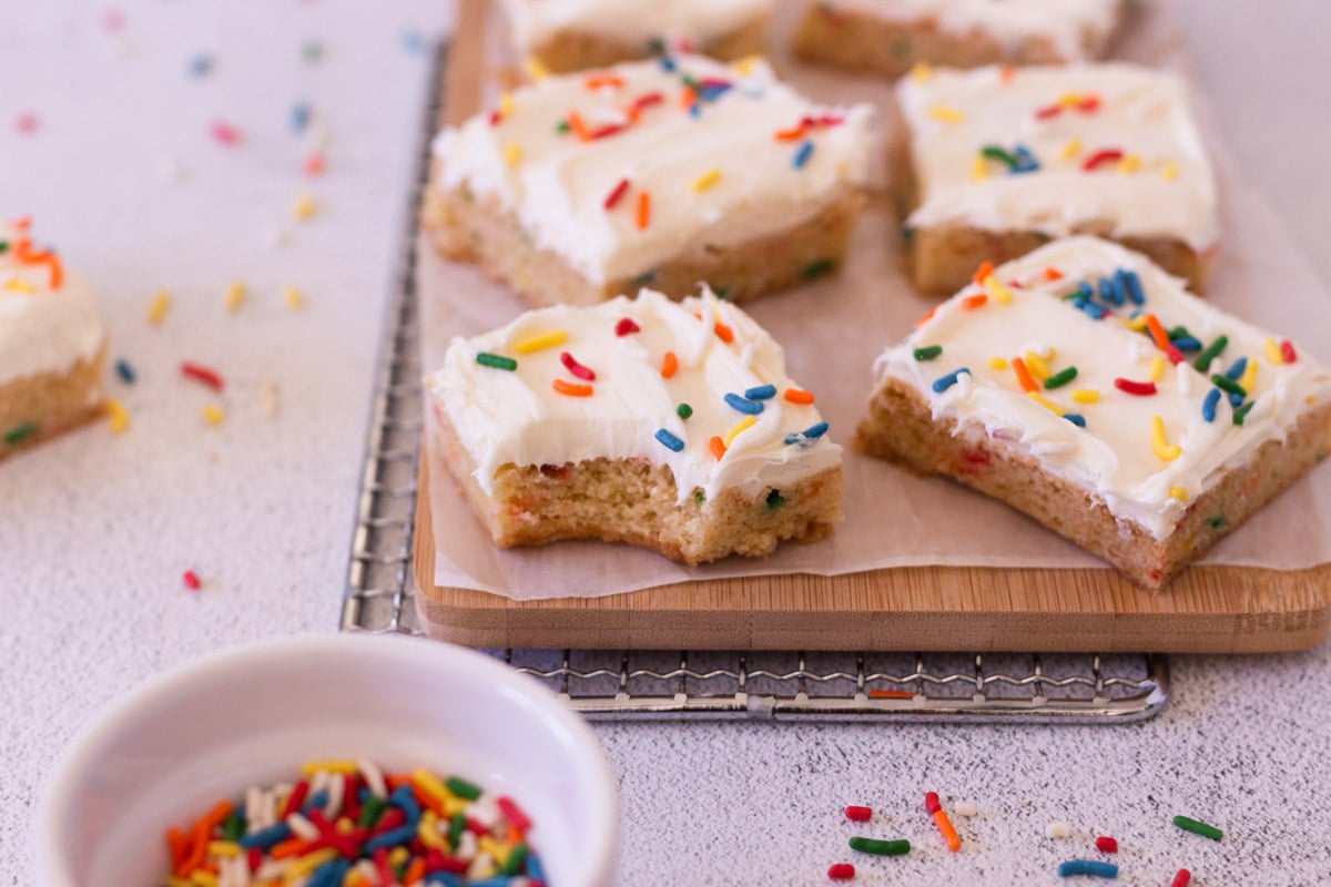 4 squares of funfetti cake mix cookie bars on a wooden cutting board, surrounded by rainbow sprinkles. 